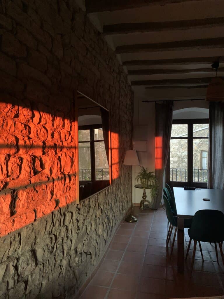 Golden-hour sunlight reflecting off the stone wall inside Casa Pardal's dining and coworking room with rustic beams and large windows.