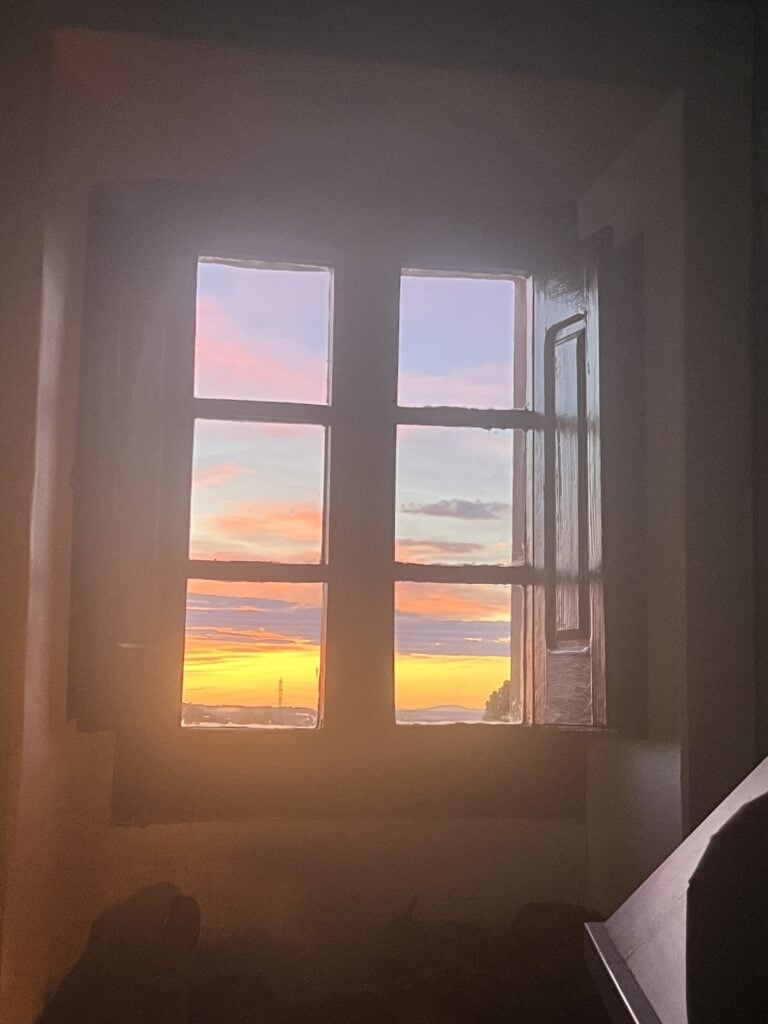 Sunset view through a rustic wooden window at Casa Pardal in Catalonia, with pastel skies and golden light filling the room. 