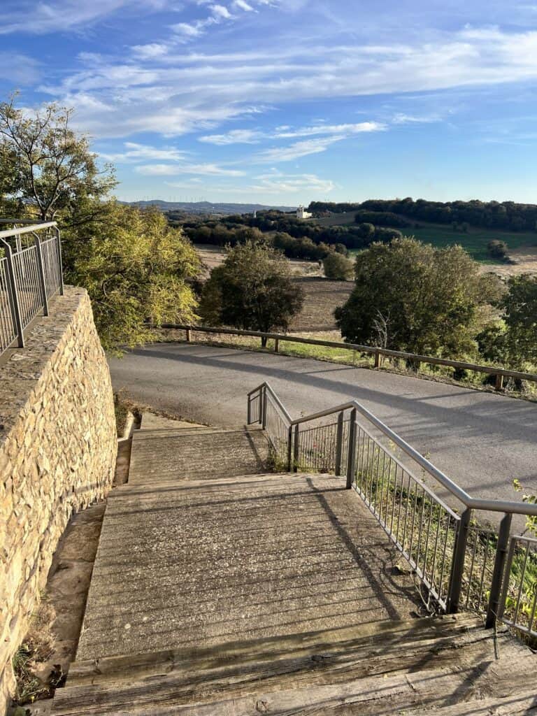 Wooden steps at Casa Pardal overlooking the rural Catalonia landscape, with open fields, trees, and hills under a bright blue sky.