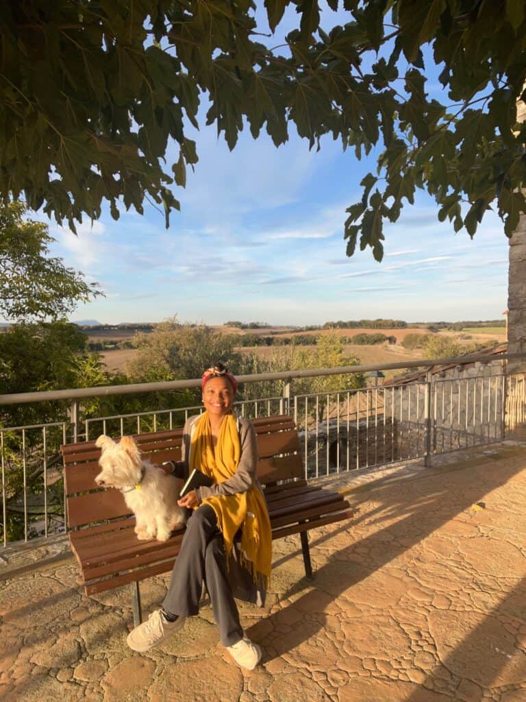 Woman journaling on a bench with Nelly the dog at Casa Pardal, overlooking the Catalonia countryside in warm afternoon light. 